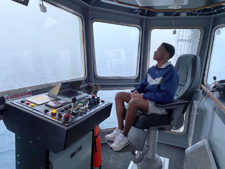 A man sitting in a chair in front of a control panel in a cockpit, looking out over the sea.