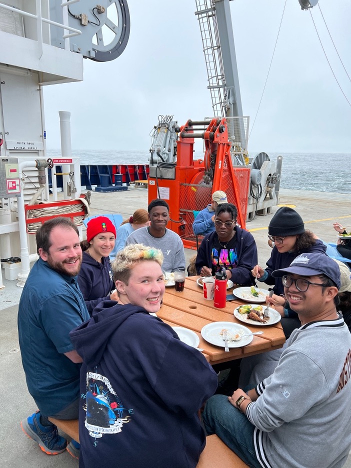 A group of students sit around a picnic table on the deck of a ship eating a meal and smiling. 