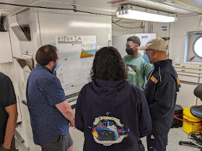 Three students standing in front of a poster presentation titled "Hypoxia," explaining the work to a man looking at the poster. 