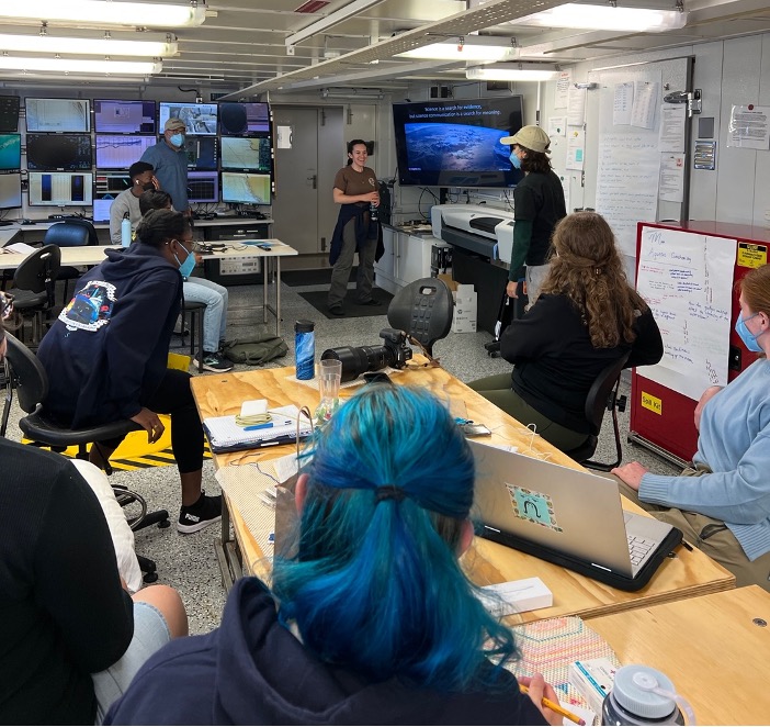 A group of people sitting in a science lab listening to a woman give a lecture. The slide has a picture of Earth on it. 