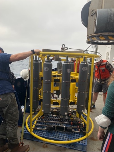 A cylindrical yellow frame, about 1.5m tall, with a dozen grey cylinders arranged inside in a circle. Students are assembling the contraption on the deck of the R/V Sally Ride wearing safety vests and hard hats.