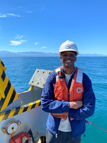 A man wearing a safety vest and hard hat, standing on the stern of a ship with the sea behind him and mountains on the horizon.