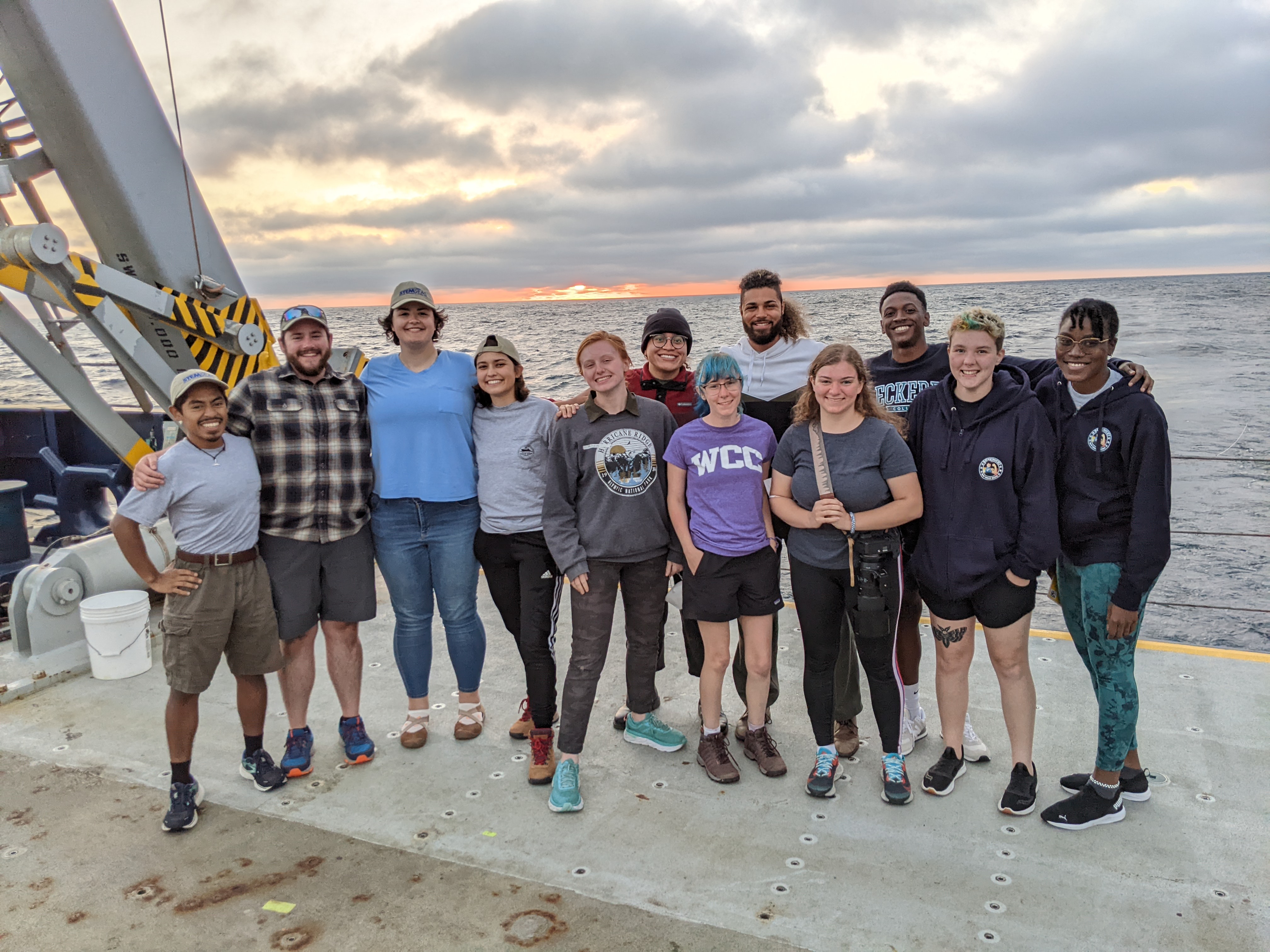 A group of 12 students standing on the fantail (stern main deck) of the R/V Sally Ride, with a sunset at sea behind them.