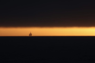 Dark, low-hanging clouds come almost all the way down to the ocean horizon, leaving a thin bar of golden sunset light with a small ship sailing on the horizon.