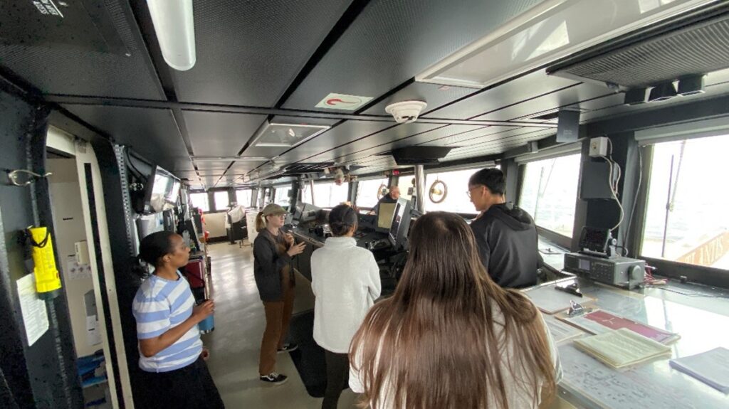 A group of students gather on the bridge of a ship with large windows looking out to the sea