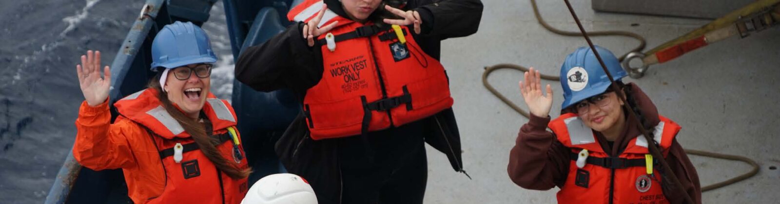 A group of scientists in red life vests and hard hats pose on the deck of a ship