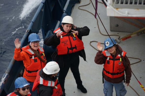 A group of scientists in red life vests and hard hats pose on the deck of a ship