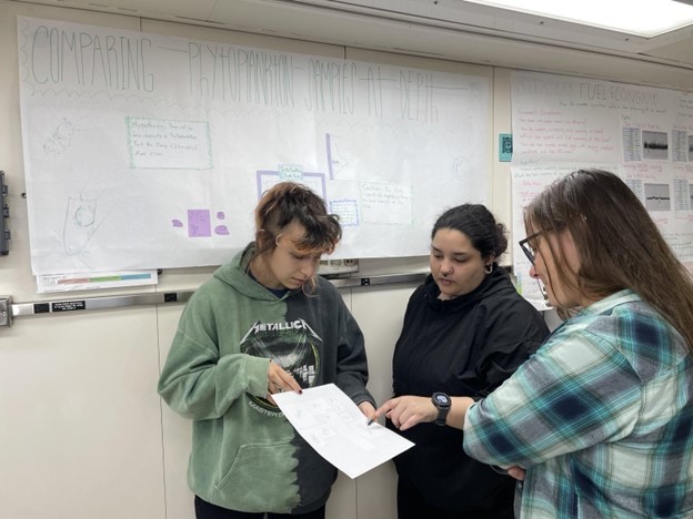 Two students point to a document to an onlooker with a scientific poster in the background