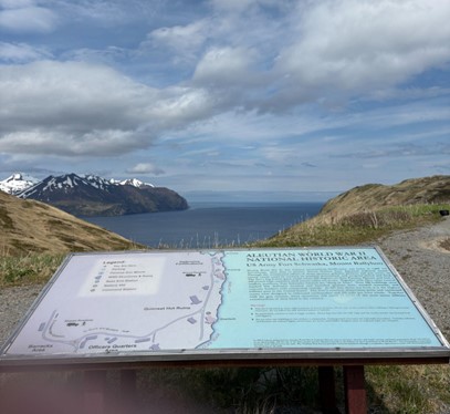 An informative sign about the location is in the foreground with a bay and snowy mountains in the background