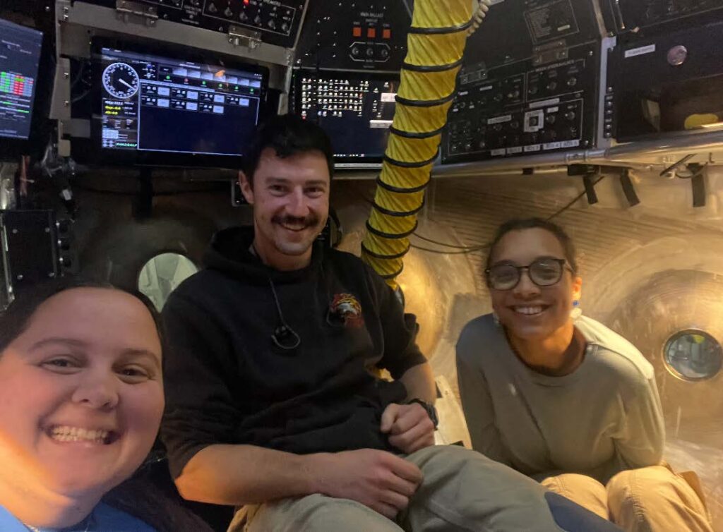Three scientists inside the Alvin bubble with computer screens and a yellow vacuum tube in the background