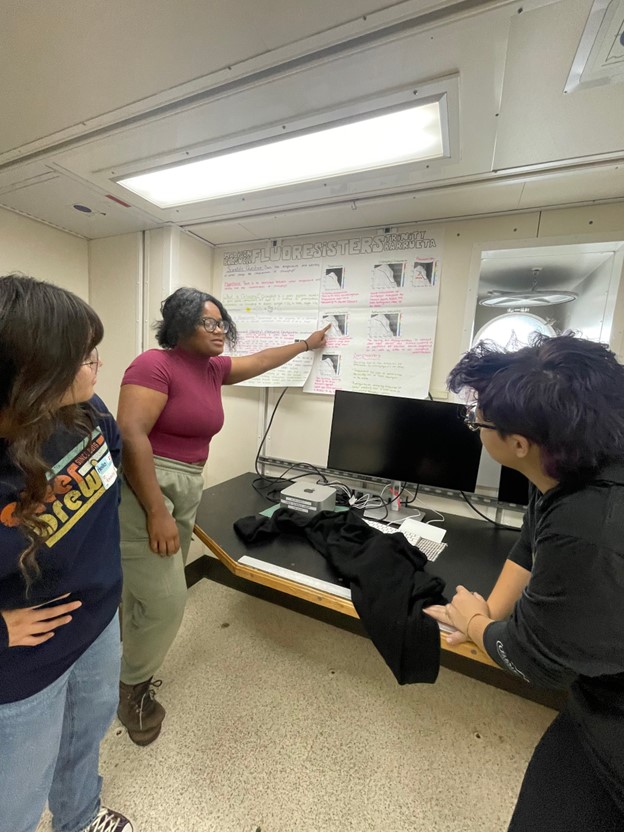 A student points at a scientific poster while two others learn from her