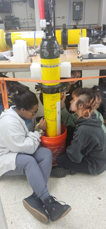 Three students sign their names on a yellow cylinder of science equipment
