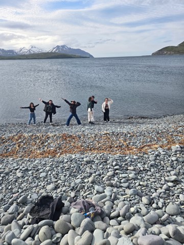 Five scientists pose by the water of a rocky beach with snowy mountains in the distance