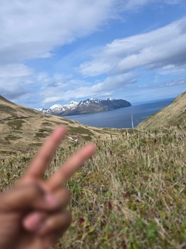 someone holds up a peace sign with a grassy valley opening up into the ocean in the background