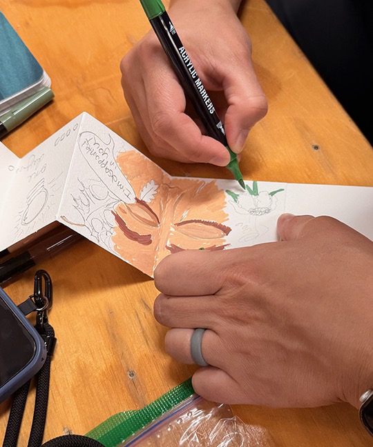Cristina's hands working on her story. a person's face is coming into view on page 1 with text bubbles. One bubble says "I'm disappointed in you!"